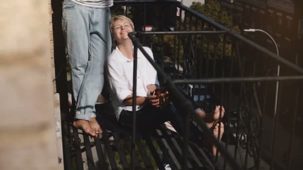Hermosa pareja multiétnica disfrutando de la mañana con café, sonriéndose mutuamente en un pequeño balcón soleado en Nueva York . — Vídeo de stock