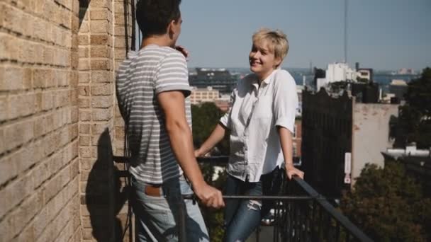 Young man and woman standing together, enjoying talking and flirting at a small sunny balcony with lovely New York view. — Stock Video