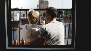 Mutlu genç arkadaşlar birlikte küçük bir New York balkon oturmak, daire penceresinden app alışveriş smartphone kullanarak görüntüleme.
