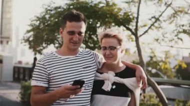 Ağır çekim bir parlak yaz gününde smartphone kullanarak İspanyol delikanlı ve Avrupa kadın yürüyüş sarılma, gülümseyen mutlu.