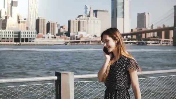 Feliz relajado sonriente chica caucásica con el pelo largo caminando a lo largo de Manhattan vista del horizonte hablando por teléfono en un día soleado — Vídeo de stock