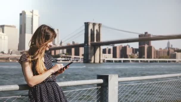 Hermosa chica caucásica sonriente con el pelo largo de pie en la famosa vista del horizonte de Nueva York usando un teléfono inteligente, alejándose . — Vídeos de Stock
