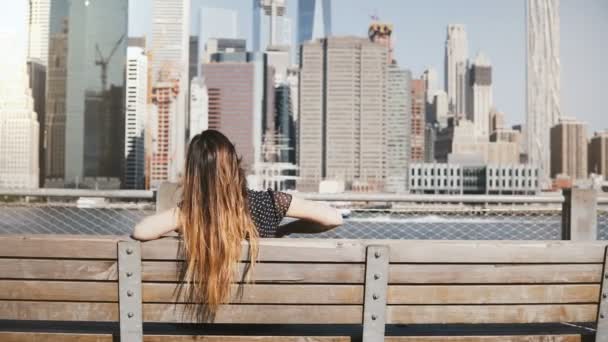 Rückansicht des glücklichen Mädchens mit langen fliegenden Haaren genießen erstaunliche manhattan Skyline Blick sitzt auf einer Bank Arme weit geöffnet. — Stockvideo