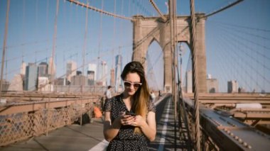 Güzel beyaz işkadını Brooklyn köprüsünde 4 k smartphone messenger app kullanarak şık güneş gözlüğü standları.