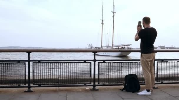 Young handsome man with backpack standing on the pier and taking photo of the motor boat on the smartphone. — Stock Video