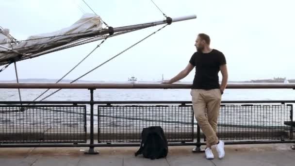 Junger gutaussehender Mann mit Rucksack steht auf der Seebrücke in der Nähe des Wassers in der Nähe der Bugspitze des Schiffes und schaut sich um. — Stockvideo