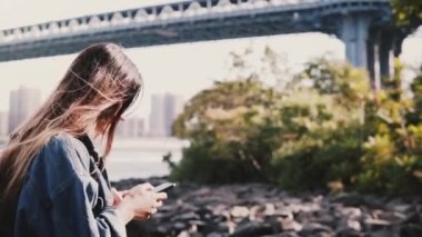 Genç gezgin kız sakin Brooklyn Köprüsü nehir banka ile smartphone ağır çekim boyunca yürüyüş sırt çantası ile yan görünüm
