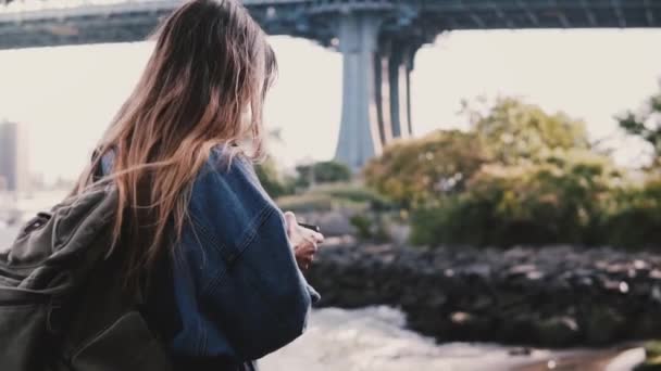 Side view of young female tourist with backpack walking along sunny Brooklyn Park river using smartphone slow motion. — Stock Video