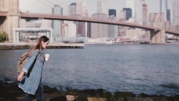 Beautiful Caucasian girl with backpack walking on stone river bank with coffee at amazing New York skyline slow motion — Stock Video