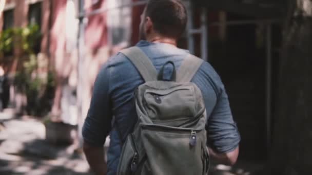 Kamera begleitet jungen entspannten Einheimischen mit Rucksack, der in New York in Zeitlupe die schattige Summer Brooklyn Street entlangläuft. — Stockvideo