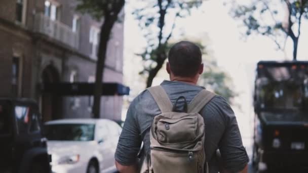 Camera follows young relaxed male freelance worker with backpack walking along shady street in Brooklyn slow motion. — Stock Video