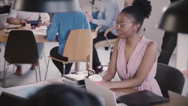 Afro-Amerikaanse vrouwelijke zakenvrouw met smartphone zitten door de office-tabel in drukke moderne naaiatelier slow motion. — Stockvideo