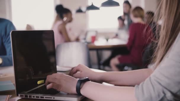 Close-up of unrecognizable young female hands with smart bracelet typing on laptop by the table, team work in background . — Stok Video