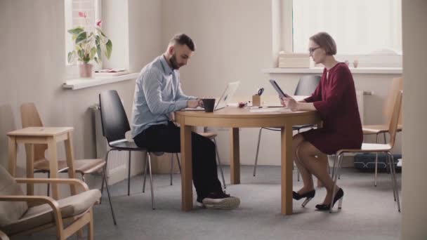 Молода жінка-бос сидить за столом через чоловіка-працівника з ноутбуком, показує йому інформацію на планшеті в сучасному світлому офісі . — стокове відео