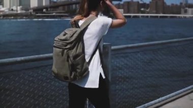 Genç kadın turist rahat Brooklyn Köprüsü'nün doğru güneşli nehir kıyısında yürüyüş zevk sırt çantası kamera takip.