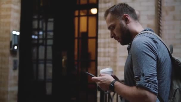Unga lokala lycklig med ryggsäck går långsamt längs skumma sommaren stadsgata med kaffe med smartphone-app. — Stockvideo