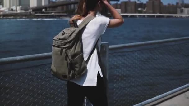 Camera follows young female tourist with backpack enjoying relaxed walking towards Brooklyn Bridge on sunny river bank. — Stock Video