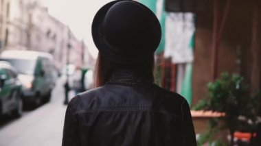 Kamera genç kadın moda blogger eski deri ceketi ve şık şapka şehir sokak ağır çekim yürüyüş izler. Mutlu yerel serbest çalışan güzel sokakları keşfetmek keyif.