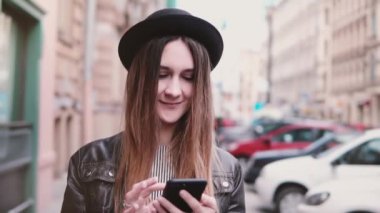 Gülümseyen genç Avrupa kadın şık şapka şehir sokak ağır çekim dört gözle bir smartphone ile yürüyüş içinde.