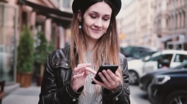 Şık şapka app ağır çekim alışveriş smartphone kullanarak bir cadde boyunca yürüyüş içinde mutlu beyaz kadın blogger gülümseyen.