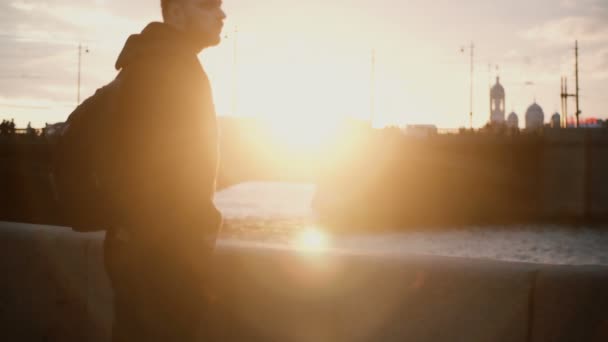 Joven hombre con estilo de pie en el terraplén cerca del río y disfrutar de la hermosa vista al atardecer. Hombre caminando por la noche . — Vídeo de stock