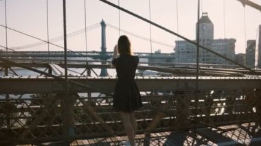 Güzel beyaz bir kadın Brooklyn Köprüsü, New York boyunca yürüyor, durur ve smartphone fotoğraf doğal görünümün 4 k alır.