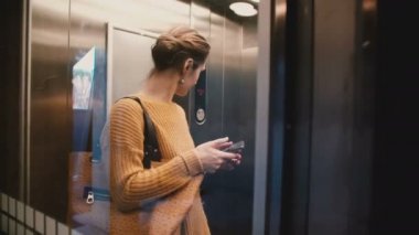 Çekici bir görünümünü meşgul beyaz bir kadın smartphone hareket eden yazıhane app kullanarak şeffaf asansöre binme vurguladı.