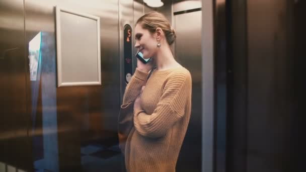 Une vue de jeune femme européenne détendue montant dans un ascenseur mural en verre transparent parlant au téléphone et souriant . — Video