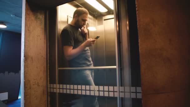 Joven hombre casual feliz montando ascensor de cristal transparente, la puerta se abre y sale con la aplicación de compras de teléfonos inteligentes . — Vídeos de Stock