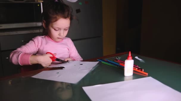 Menina europeia pré-escolar bonita em suéter rosa sentado à mesa em casa sozinho formas de corte de papel . — Vídeo de Stock