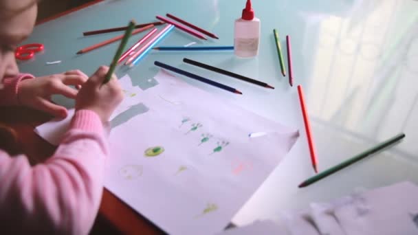 Vue du dessus caméra coulissante à gauche sur une petite fille caucasienne dessin d'enfant sur papier avec divers crayons de couleur à une table . — Video