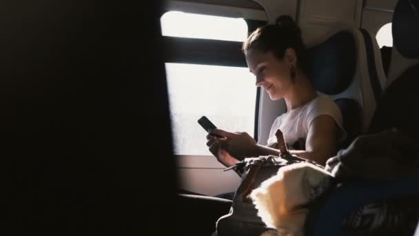 Belle fille heureuse caucasienne communiquant avec des amis en utilisant un smartphone et souriant tout en voyageant dans le train . — Video