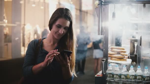 Feliz hermosa bloguera caucásica usando la aplicación de compras de teléfonos inteligentes cerca de camión de comida callejera con pretzels en Nueva York . — Vídeo de stock