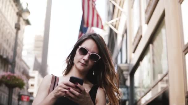 Счастливая белая деловая женщина в солнцезащитных очках с помощью приложения для смартфонов на улице Нью-Йорка с флагами США замедленной съемки — стоковое видео