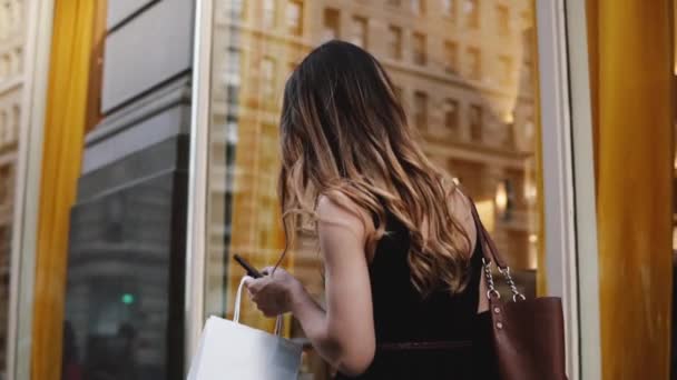 La caméra suit une jeune blogueuse beauté portant des sacs à provisions dans les deux mains marchant le long de la rue au ralenti . — Video