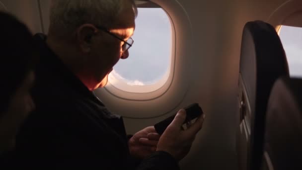 Gelukkig senior mannelijke vliegtuig passagier controleren van zijn smartphone, genieten van het uitzicht tijdens de vlucht, zittend in het venster. — Stockvideo