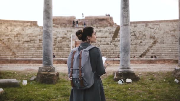 Achteraanzicht van mooie toeristische meisje met rugzak staan voor oude amfitheater pijlers in Ostia, Italië. — Stockvideo