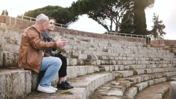 Gelukkig Europese senior man en jonge vrouw zittend op de ruïnes van oude amfitheater in Ostia, Italië met een kaart en een smartphone. — Stockvideo