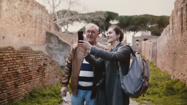 Pria senior yang gembira dan wanita muda Eropa yang tersenyum berfoto selfie di dekat reruntuhan tua di Ostia, Italia dalam perjalanan liburan . — Stok Video