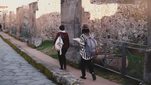 Cámara sigue a dos mujeres con un mapa caminando a lo largo de antiguas ruinas y calles de Pompeya, Italia en vacaciones de viaje . — Vídeo de stock
