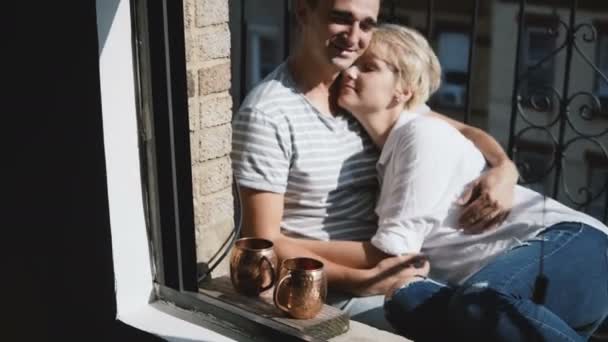Hermosa joven pareja romántica multiétnica sentados juntos, abrazando y hablando en un pequeño balcón soleado apartamento . — Vídeos de Stock