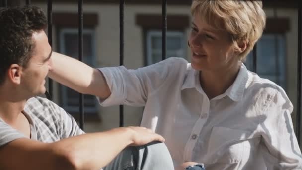 Krásná mladá mnohonárodnostní romantický pár seděli v malé slunný balkon pohledu a mluvit k sobě navzájem. — Stock video