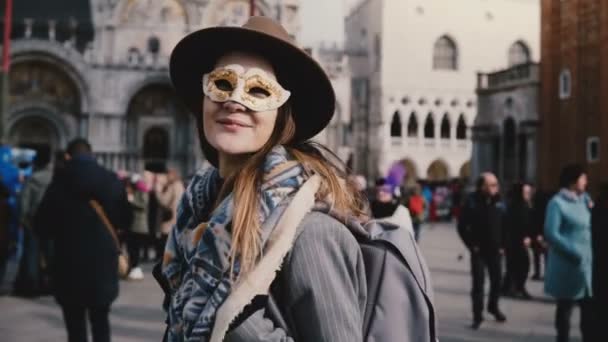 Porträt einer glücklichen Frau mit langen Haaren, die eine weiße Karnevalsmaske auf dem venezianischen Stadtplatz trägt, Italien bläst Küsse.