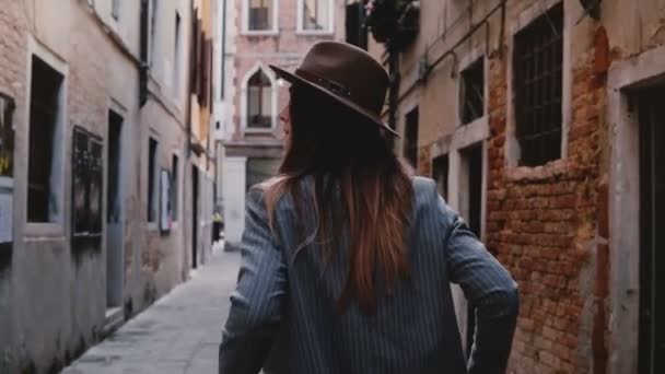 Indietro vista di fiduciosa giovane donna d'affari elegante in cappello e tuta passeggiando lungo la stretta strada antica a Venezia, Italia . — Video Stock