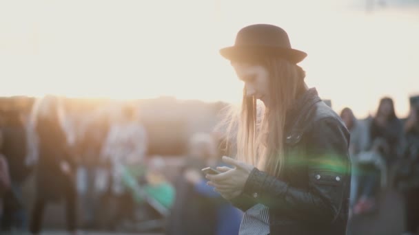 Joven hermosa mujer de pie en la calle llena de gente y el uso de teléfonos inteligentes. Elegante hembra en la ciudad al atardecer . — Vídeos de Stock