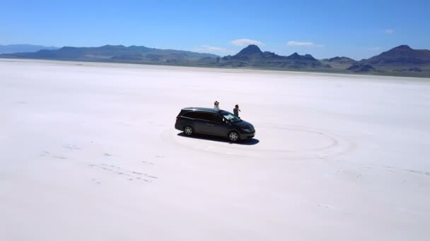ミニバン車ボンネビル塩湖砂漠の真ん中で腕と興奮してカップルの息をのむの空中ショットを開く. — ストック動画