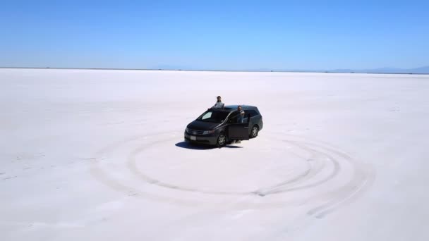 Drone flying close around excited couple at silver minivan car in the middle of amazing Bonneville salt desert road trip — Stock Video