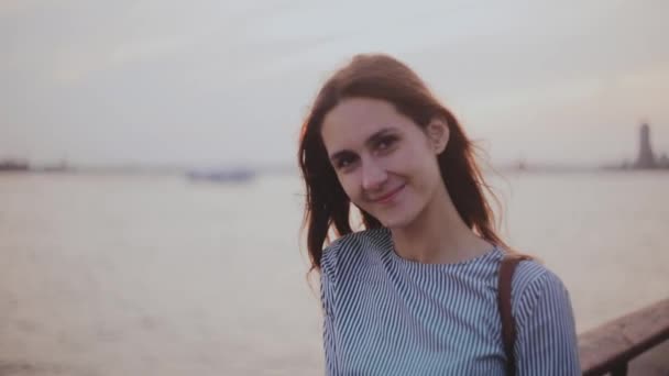 Portret van gelukkig jong Europees meisje poseren, op zoek naar camera met haar waait in de wind op zonsondergang rivier strand. — Stockvideo