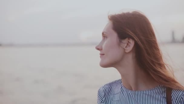 Retrato de la joven europea reflexiva feliz con el pelo soplando en el viento y sonrisa tranquila en la playa del río puesta del sol . — Vídeo de stock