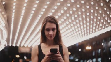 Mutlu güzel gülümseyen yerel kadın smartphone messenger, sola bakıyor kullanarak şaşırtıcı Chicago tiyatroda ayakta.